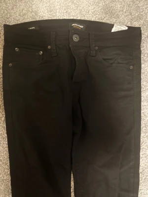 Jack & Jones slim jeans - Snygga svarta jeans från Jack & Jones med klassisk femficksdesign och smal passform. Modellen är slim/Glenn och har en enkel look med svarta nitar och knapp. Perfekta för dig som gillar stilrena och tajta jeans.