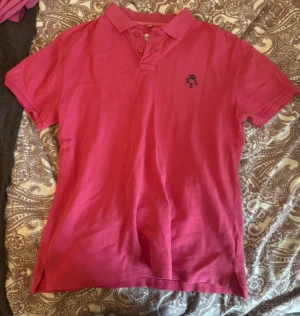 Rosa pikétröja KSSS med krage - Snygg rosa pikétröja från KSSS med klassisk krage och knappar framtill. Tröjan har broderad logga på bröstet och texten KSSS på ryggen. Tillverkad i mjuk bomull, perfekt för en avslappnad och sportig look.