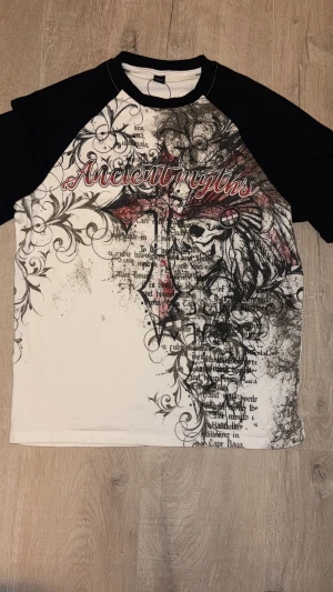  t-shirt med tryck - Snygg t-shirt med svart-vita kontraster och coolt grafiskt tryck framtill med texten 'Ancient Myths' i rött. Kortärmad med svart krage och ärmar, vit bas och detaljerade mönster i svart och rött. Perfekt för dig som gillar unika prints och streetstyle.