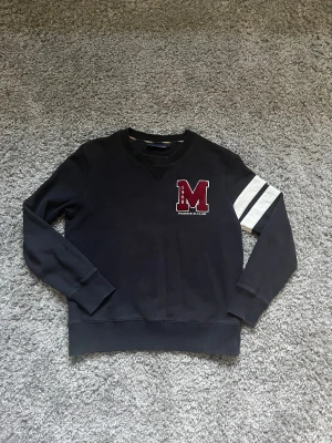 Svart sweatshirt Morris R. Club S - Svart sweatshirt från Morris R. Club i storlek S med broderad röd M-logga på bröstet och vita ränder på ena ärmen. Tröjan har rund hals och ribbade muddar vid ärmslut och nederkant. Perfekt för dig som gillar college-stil.