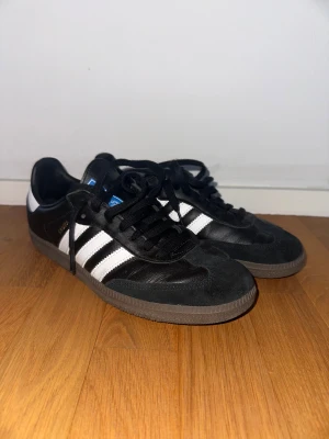 Adidas Samba svarta sneakers - Klassiska Adidas Samba sneakers i svart skinn och mocka med vita ränder och brun sula. Ikonisk retrostil med snörning och rund tå, perfekt för dig som gillar streetwear och sportig vibe.