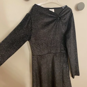 Glittrig svart kortklänning från Zara - Svart kortklänning från Zara med glittriga prickar över hela tyget. Klänningen har lång ärm och en snygg detalj med ringning framtill vid halsen. Materialet är mjukt och stretchigt, perfekt för dig som gillar att sticka ut med lite extra glitter.