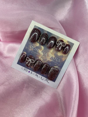 GlamGlow (S); ExtraShortOval  - Säljer ett set handgjorda press-on naglar i storlek S: 15 mm, 11,5 mm, 12 mm, 11 mm, 9 mm. Naglarna är vinröda med silverfärgadglitter och har en extra kort oval form. Två av naglarna har en söt silverrosett som 3D-dekoration. Perfekt för dig som vill ha unika och snygga naglar utan att gå till salong.