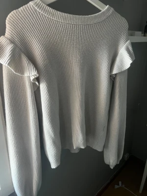 Vit stickad tröja med volangdetaljer - Säljer en vit stickad tröja med snygga volangdetaljer på axlarna. Tröjan har en ribbad struktur och långa ärmar, perfekt för dig som gillar en enkel men ändå trendig look. Passar dig som vill ha något mjukt och bekvämt till garderoben.