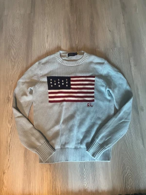  Ralph Lauren tröja - Snygg ljusblå stickad tröja från Polo Ralph Lauren med amerikansk flagga i rött, vitt och marinblått på bröstet. RL-broderi under flaggan. Tröjan har rund hals, ribbade muddar och lång ärm. Perfekt för dig som gillar klassisk och cool stil.