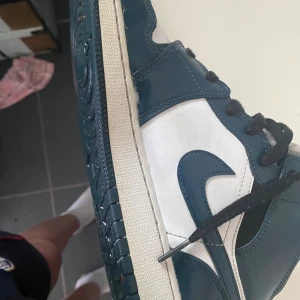 Nike Air Jordan 1 Mid blå/vit - Säljer ett par Nike Air Jordan 1 Mid i blått och vitt med svarta skosnören. Skorna har klassisk Jordan-design med rund tå, snörning och vit sula. Ovandelen är i skinn och syntet, vilket ger en clean och sportig look. Perfekta för dig som vill sticka ut med stil.