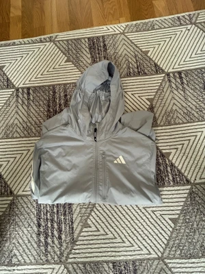 Grå vindjacka från Adidas med huva - Snygg grå vindjacka från Adidas med huva och dragkedja framtill. Jackan har reflekterande detaljer, lätt material och är perfekt för löpning. Adidas-logga på bröstet och texten WIND.RDY på ärmen. Sportig och stilren design. Och ett litet hål brevid bröstfickan