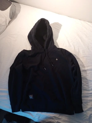 Svart hoodie från Morris med huva - Svart hoodie från Morris med broderad logga på bröstet och snörning i huvan. Tröjan har lång ärm och en klassisk passform, perfekt för chill dagar. Materialet är mjuk bomull och polyester, vilket gör den skön att bära. Snygg detalj med patch nertill.