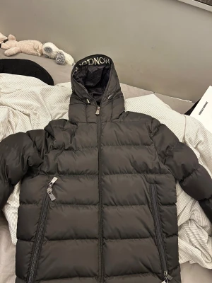 Svart Moncler pufferjacka med huva - Intressekoll! Svart pufferjacka från Moncler med huva och tydlig logga på axeln samt på insidan av jackan. Jackan har dragkedjor framtill och på fickorna. Den är i en storlek 2. Materialet är polyester och dun, perfekt för kalla dagar. Snygg och stilren look med loose passform. Den är i nyskick. Väldigt varm och skön jacka nu till vintern. Hör av dig vid frågor! 