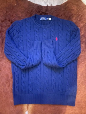 Blå kabelstickad tröja Polo Ralph Lauren - Kabelstickad tröja från Polo Ralph Lauren i en snygg blå färg med rund hals och ribbade muddar. Tröjan har lång ärm och den klassiska röda Polo-loggan broderad på bröstet. Materialet är mjuk bomull som känns skön mot huden.