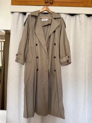 Beige trenchcoat från Calvin Klein - Stilren beige trenchcoat från Calvin Klein med klassisk dubbelknäppning, bred krage och justerbara ärmslut. Lång modell med fickor och snygga detaljer. Perfekt för dig som vill ha en tidlös och clean look.