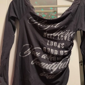 Grå långärmad topp med spets - Grå långärmad topp med bred, rundad halsringning dekorerad med spets. Tryck i silverfärg med text över hela framsidan. Materialet känns mjukt och stretchigt, perfekt för dig som gillar statement-plagg med lite edge.