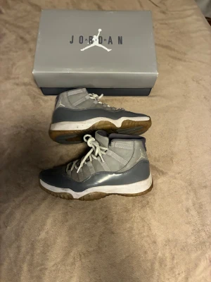 Cool grey Jordan 11 s - Säljer ett par gråa Nike Air Jordan 11 sneakers med högt skaft och vita snören. Skorna har en blandning av blankt och matt material, med vit mellansula och genomskinlig gummisula. Ikonisk Jumpman-logga på sidan och klassisk Jordan-box medföljer.