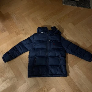 Mörkblå dunjacka från Polo Ralph Lauren - Snygg mörkblå dunjacka från Polo Ralph Lauren med röd broderad logga på bröstet. Jackan har huva, dragkedjor på fickorna och är quiltad för extra värme. Perfekt inför vintern när det är kallt. Passar 14 till 16 år, men jag skulle rekommendera för lite yngre pågrund av att märket gör lite mindre storlekar i barn.