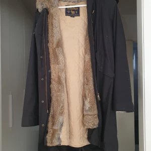 Svart parkajacka med pälsfoder från Woolrich - Säljer en svart parkajacka från Woolrich med mysigt pälsfoder och huva. Jackan har långa ärmar och stängs med både dragkedja och knappar. Perfekt för kalla dagar med sin quiltade insida och stilrena design.