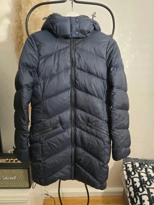 Mörkblå lång dunjacka från Gant - Snygg mörkblå dunjacka från Gant med huva och dragkedja framtill. Jackan är lång, har två snedställda fickor med dragkedja och ett Gant-märke på ärmen. Perfekt för kalla dagar och har en stilren, quiltad design. Finns en liten knappt märkbar fläck framtill annars i jättefint skick!