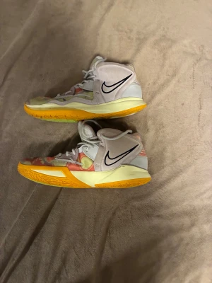 Nike Kyrie infinity basket sneakers med mönster - Säljer ett par Nike Kyrie basket sneakers med coolt abstrakt mönster i rött, gult och lila. Skorna har högt skaft, snörning och tydlig Nike-logga på sidan. Sulan är gul och ovandelen är i syntetmaterial med mesh och mockadetaljer. Perfekta för dig som vill sticka ut på planen.
