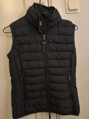 Svart dunväst från Parajumpers - Svart quiltad dunväst från Parajumpers med hög krage och dragkedja framtill. Västen har två sidofickor med dragkedja och en insida i ljusgrått med en säkerhetsficka markerad med orange detalj. Super lightweight-modell, perfekt för lager-på-lager.