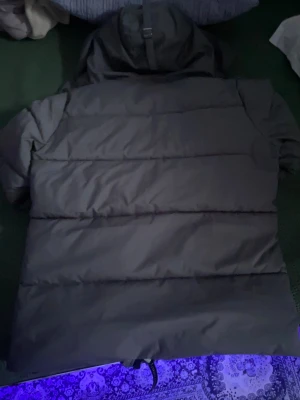 Svart pufferjacka från Canada Goose - Säljer en svart pufferjacka från Canada Goose med huva och snygga detaljer. Jackan har dragkedja och knappar framtill, två sidofickor och ett klassiskt märke på ärmen. Perfekt för kalla dagar med sin tjocka och quiltade design.