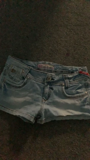 Ljusblå jeansshorts från Denim - Säljer ett par ljusblå jeansshorts från Denim med snygga detaljer på bakfickorna, inklusive nitar och broderi. Klassisk femficksmodell med knapp och dragkedja framtill. Perfekta för sommaren och har en riktigt fräsch vibe. Har tyvärr ett hål på innersta sidan av benet men det syns inte när man har dom på. Om du är intresserad kan jag skicka bild. Dom är även i storlek m men skulle säga att dom passar som s.