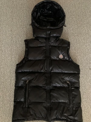 Svart dunväst från Moncler med huva - Svart dunväst från Moncler med glansig finish, avtagbar huva och två fickor med dragkedja. Klassisk Moncler-logga på bröstet och snygga tryckknappar. Perfekt för lager-på-lager och streetstyle. Insidan har en unik serietidningsetikett.