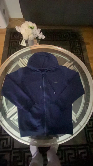Marinblå Lacoste hoodie  - Säljer en marinblå hoodie från Lacoste med dragkedja framtill och snörning vid huvan. Tröjan har långärmade ärmar och en liten broderad logga på bröstet. Perfekt för chill dagar och enkel att matcha med jeans eller joggers.