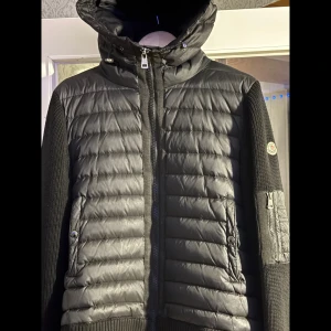 Svart Moncler pufferjacka med huva - Säljer en svart pufferjacka från Moncler med stickade ärmar och vadderad front. Jackan har huva, dragkedja framtill och Moncler-logga på ärmen. Snygg och stilren design med ribbade muddar och praktiska fickor med knappstängning och passar M också 