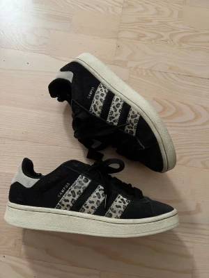 Adidas Campus sneakers med leopardmönster - Svarta Adidas Campus sneakers med beige och svart leopardmönster på de klassiska tre ränderna. Skorna har rund tå, platt sula och snörning. Ovandelen är i mocka och detaljer i syntet. Perfekt för dig som vill sticka ut med coola detaljer. 