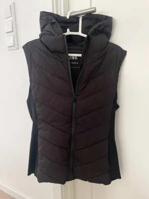 Svart dunväst med huva från ZARA - Svart dunväst från ZARA med snygg quiltad design och stor, mysig huva. Västen har dragkedja framtill och är perfekt för lager-på-lager. Materialet är polyester och passformen är normal. Stilren look som funkar till många outfits.