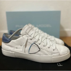 Philippe Model vita sneakers med blå detalj - Snygga vita sneakers från Philippe Model med blå häl och coolt broderat mönster på sidan. Skorna har rund tå, platt sula och klassisk snörning. Tillverkade i skinn för en lyxig känsla och har en stilren, modern look som passar till jeans eller shorts. Box och gympapåse medfölje, men boxen är rätt sliten.