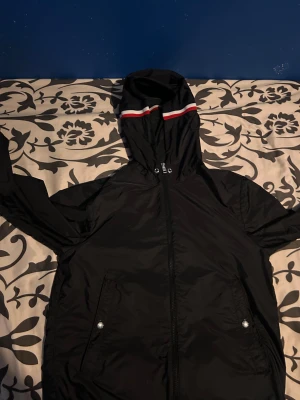 Svart vindjacka från Moncler - Svart vindjacka från Moncler med huva som har vita och röda ränder. Jackan har dragkedja framtill, två fickor med tryckknappar och ett Moncler-märke på ärmen. Materialet är lätt och syntetiskt, perfekt för blåsiga dagar.