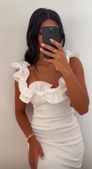 Vit kortklänning med volang - Supersnygg vit kortklänning med breda volangdetaljer runt axlar och urringning. Klänningen är ärmlös och har en figurnära passform med ribbad struktur. Perfekt för dig som vill ha en trendig och feminin look med extra statement vid axlarna.