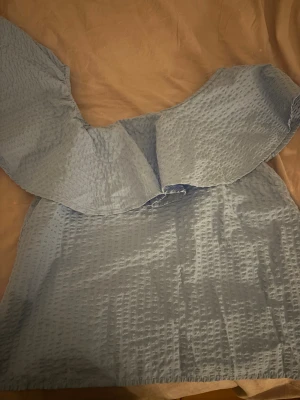 Ljusblå offshoulder blus med volang - Säljer en ljusblå blus med offshoulder-design och bred volang över axlarna. Toppen har en cool textur med rutmönstrad struktur i tyget och är ärmlös. Perfekt för dig som gillar att sticka ut med en unik och trendig look.