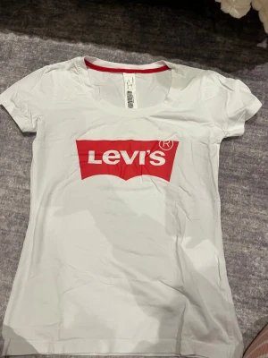 Vit Levi's t-shirt med röd logga - Vit t-shirt från Levi's med klassisk röd logga framtill. T-shirten har rund halsringning med röd kant på insidan och är kortärmad. Tillverkad i mjuk bomull för en skön känsla och enkel stil.