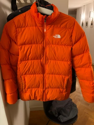 Orange dunjacka från The North Face - Säljer en riktigt fet orange dunjacka från The North Face. Jackan har en klassisk pufferstil med hög krage, dragkedja framtill och logga på både bröstet och ryggen. Perfekt för kalla dagar när du vill sticka ut med färg. Snygg och funktionell design. Jackan är även reversibel och kan bli svart med orange dragkedja.