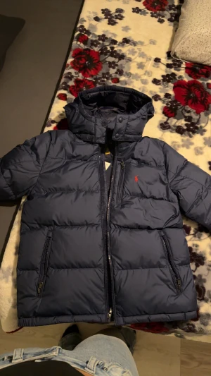 Mörkblå dunjacka från Polo Ralph Lauren - Säljer en helt ny mörkblå vinterjacka med prislapp kvar, köpt från kidsbrandstore. Jackan är i storlek L (14-16 år) och har inte använts en enda gång. Den är varmt, lätt och perfekt för vintern, med avtagbar huva, flera praktiska dragkedjefickor och en stilren broderad röd logo på bröstet. På den fjärde bilden är det en Qr kod som tar dig direkt till Polo Ralph Lauren hemsidan. Faktura och beställningsorder finns tillgänglig. Priset kan diskuteras! 