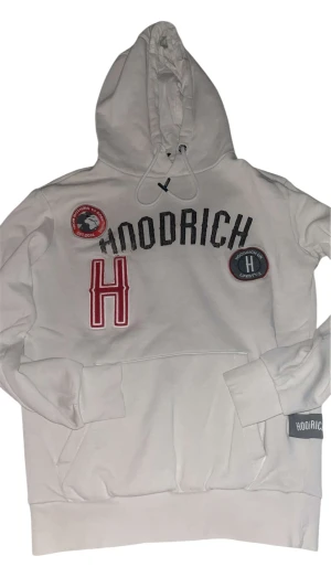 Vit hoodie från Hoodrich med patchar - Säljer en vit hoodie från Hoodrich med broderade patchar och tryck på bröstet. Tröjan har huva med snörning, stor ficka framtill och ribbade muddar. Hoodrich-logga på ärmen och coola detaljer som ger streetkänsla.