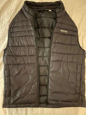 Svart dunväst från Jack & Jones XS - Snygg svart dunväst från Jack & Jones i storlek XS. Västen har quiltad design, diskret logga på bröstet och dragkedja framtill. Perfekt att slänga över en hoodie för extra värme. Materialet är syntet och polyester, vilket gör den lätt och smidig.