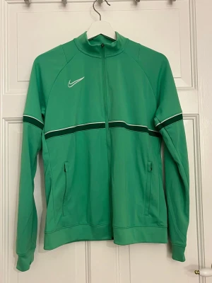 Grön Nike Dri-Fit ziptröja M - Nike Dri-Fit ziptröja i grön färg med vita och mörkgröna detaljer. Tröjan har hel dragkedja, ståkrage och två fickor med dragkedja. Swoosh-logga på bröstet och sportig design, perfekt för träning eller chill. Materialet är lätt och andas.
