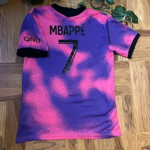 PSG Mbappé matchtröja rosa/lila - Säljer en PSG fotbollströja med Mbappé 7 på ryggen. Tröjan har ett unikt rosa och lila mönster med svarta detaljer, klubbmärke på bröstet och sponsortryck. Tillverkad i lätt och ventilerande polyester, perfekt för dig som vill sticka ut på planen.