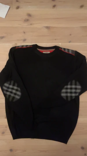 Mörkblå Burberry tröja med rutiga detaljer - Snygg mörkblå tröja från Burberry med klassiska rutiga detaljer på armbågar och axlar, samt en röd linje längs axlarna. Tröjan har rund hals och är långärmad, med diskret Burberry-logga broderad på bröstet. Perfekt för dig som gillar stilrena och unika plagg.