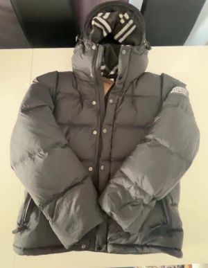 Svart pufferjacka från Burberry XL - Svart pufferjacka från Burberry London England i storlek XL men känns som L. Den är helt oanvänd. Jackan har huva, dragkedja och knappar framtill samt två sidofickor med dragkedja. Insidan har ett snyggt randigt foder i grått och vitt. Perfekt för kalla dagar med sin tjocka och fluffiga design.