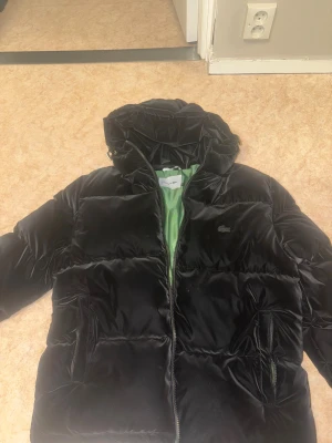 Svart pufferjacka från Lacoste - Snygg svart pufferjacka från Lacoste med broderad krokodil på bröstet och stor logga på ryggen. Jackan har huva, dragkedja och grönt foder. Perfekt för kalla dagar och riktigt clean look.