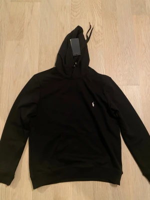 Svart hoodie från Polo Ralph Lauren - Svart hoodie från Polo Ralph Lauren med klassisk vit broderad logga på bröstet. Tröjan har huva med snörning, känguruficka och långa ärmar. Tillverkad i mjuk bomull som är skön att ha på sig. Perfekt för dig som gillar stilrena och tidlösa plagg.