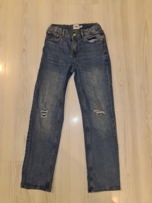 Blå jeans från 157 med slitningar - Säljer ett par klassiska blå jeans från 157 i storlek 160. Jeansen har raka ben, snygga slitningar på båda knäna och femficksmodell. De har normal passform och är tillverkade i jeansmaterial med orangea sömmar och knappstängning framtill.