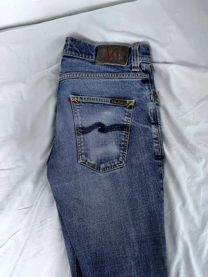 Blå jeans från Nudie Jeans Co W30 L32 - Snygga blå jeans från Nudie Jeans Co med klassisk femficksdesign och coola kontrastsömmar i orange. Jeansen har en mörkblå vågig brodering på bakfickan och läderpatch med logga. Passar dig som gillar stilrena och tidlösa jeans med schyssta detaljer.