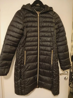 Svart lång pufferjacka MyWear Active - Svart lång pufferjacka från MyWear Active med huva och guldfärgade dragkedjor. Jackan har quiltad design, två fickor med dragkedja och elastiska muddar vid ärmslut. Perfekt för kalla dagar och har en snygg, sportig look.