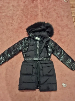 Svart pufferjacka Calvin Klein Jeans - Svart pufferjacka från Calvin Klein Jeans med glansig finish, huva med fuskpäls och justerbart bälte med logotyp. Jackan har stora fickor framtill, dragkedja och lång ärm. Perfekt för kalla dagar med sin stilrena look och coola detaljer.