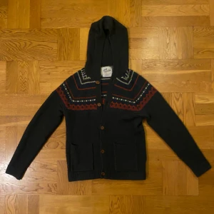 Mörkblå stickad hoodie med mönster - Mysig mörkblå stickad hoodie med snyggt mönster i rött, blått och vitt över axlar och bröst. Tröjan har huva, knappar framtill och två praktiska fickor. Perfekt för kalla dagar, antingen som hoodie eller som överlager med en dunväst. 