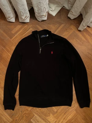 Svart half zip tröja Polo Ralph Lauren - Svart långärmad tröja från Polo Ralph Lauren med klassisk röd broderad logga på bröstet. Tröjan har en half zip-dragkedja vid halsen och är tillverkad i ett mjukt bomullsmaterial. Perfekt för lager-på-lager och stilren look.